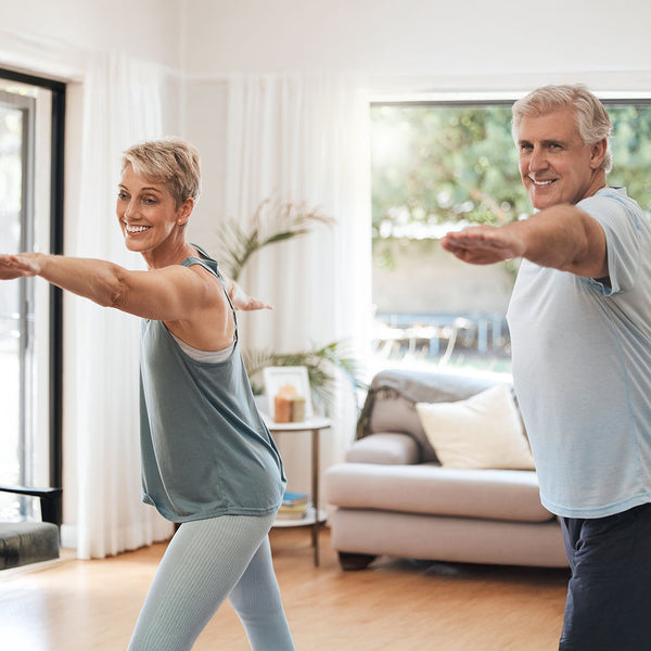 thema rueckenfit - man sieht ein paar im wohnzimmer, sie tragen beide sportliche kleidung und führen eine übung aus bei der sie jeweils den linken arm nach vorne strecken und sich auch leicht nach vorn beugen. beide lächeln zufrieden.