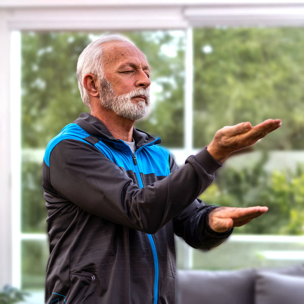 thema tai-chi - man sieht einen mann in seinem wohnzimmer. er trägt eine sportliche jacke, und führt mit geschlossenen augen eine tai chi übung durch.