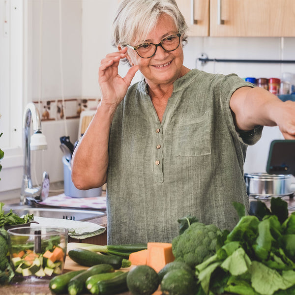 thema vegane-ernaehrung - man sieht eine frau mit grauen kurzen haaren und brille in ihrer küche. auf dem brett vor ihr sind unzähliche grüne gemüsesorten und ein mixer  ausgebreitet. sie lächelt und schaut sich ein ernährungsvideo auf dem handy an